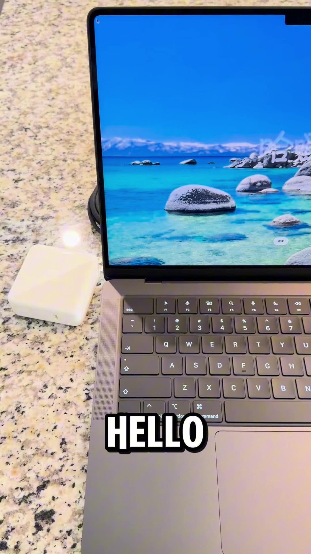 An open space-gray MacBook Pro on a speckled granite counter, its screen showing a bright alpine lake, a white power adapter beside it, and the word HELLO across the keyboard.